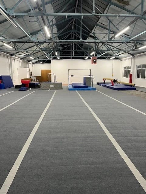 Large indoor gym hall with light grey carpet roll-out mats laid in lanes toward gymnastics apparatus at the far end.