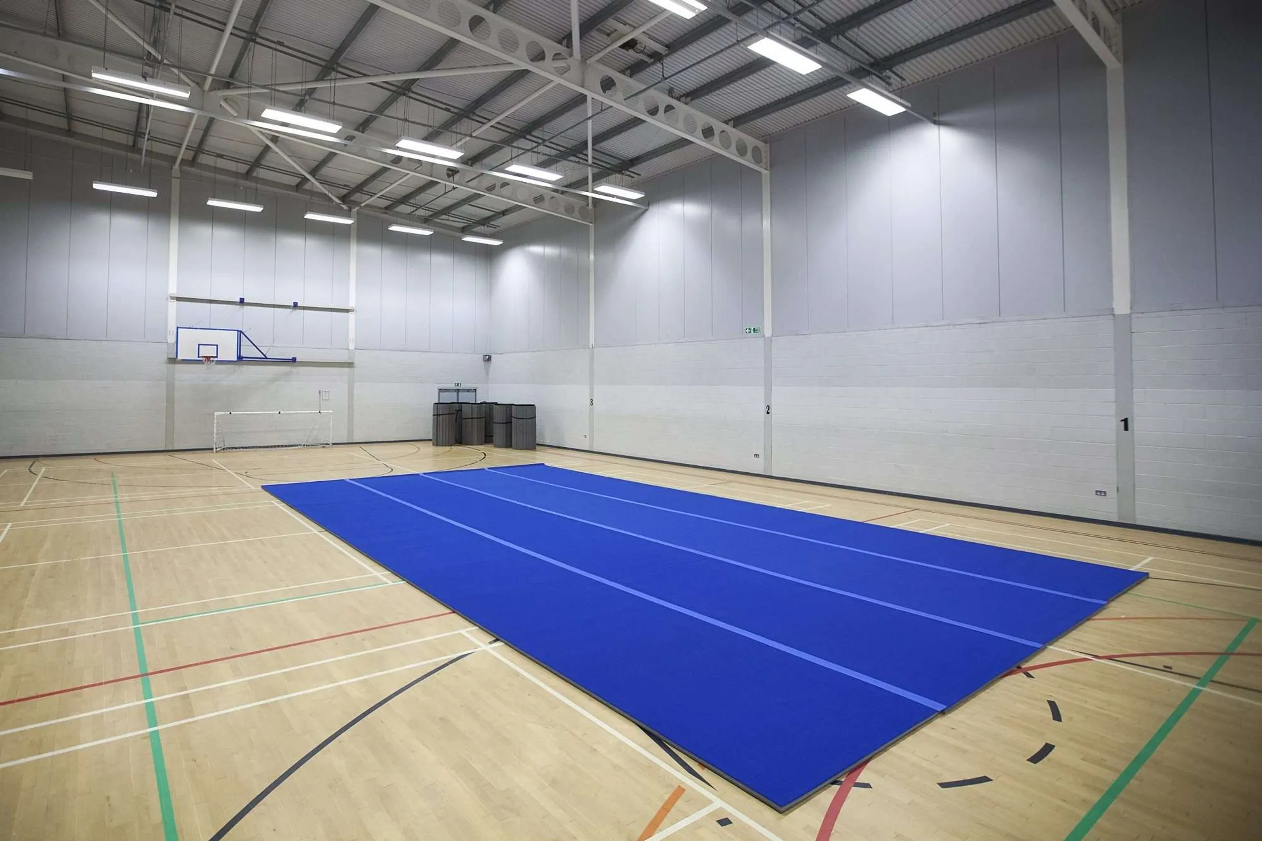 Blue roll-out carpet mat laid across the center of a large indoor sports hall with a basketball hoop and wooden floor.